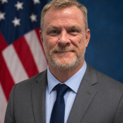 Man in business attire stands in front of American flag, smiling.