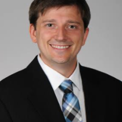 Man in black suit and blue plaid tie smiling for the camera.