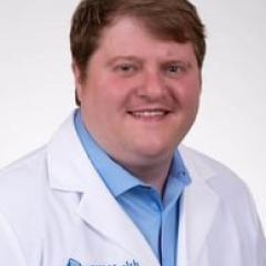 A man in a white lab coat and blue shirt smiles at the camera.