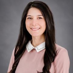 Woman with long black hair wearing pink sweater smiling against gray background.