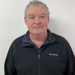 Man with gray hair, wearing black jacket and blue shirt, smiling against white background.