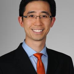 Portrait of a man in a black suit and blue shirt, wearing a red tie and glasses.