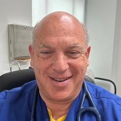 A smiling bald man in blue scrubs with a stethoscope around his neck.