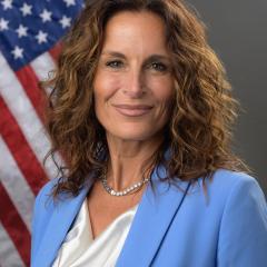 Woman with curly hair wearing a blue blazer and white blouse, standing in front of an American flag.