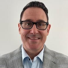 Man in gray plaid blazer and blue shirt, wearing glasses, smiling at the camera.
