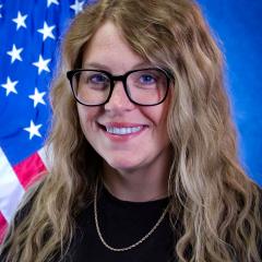 A woman with long blonde hair and glasses stands in front of an American flag.