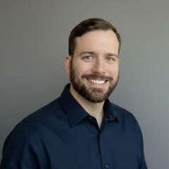 Man with short brown hair and goatee, wearing a navy button-up shirt, smiling against a gray background.