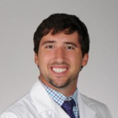 A man in a white lab coat and blue tie smiles.