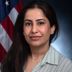 Woman with long dark hair wearing a light blue shirt, in front of the US flag.