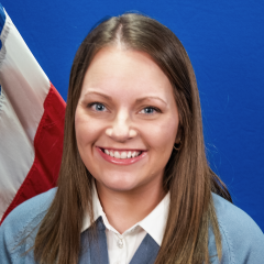 Woman in blue sweater and white shirt smiling with flag behind her.