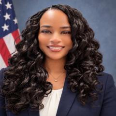 Woman with curly black hair smiling in front of American flag.