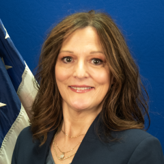 A woman in a dark blazer and gray shirt smiles in front of a flag.