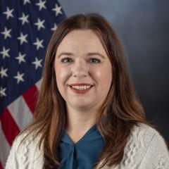 Woman in blue shirt and white cardigan smiling with American flag behind her.