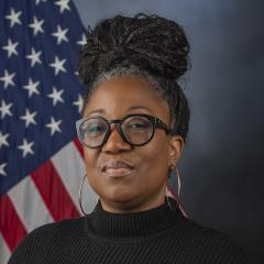 Woman in black turtleneck and glasses stands in front of American flag.