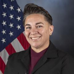 Woman with short hair, wearing a maroon shirt and black blazer, smiles in front of American flag.
