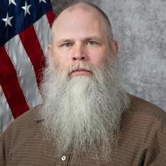 Man with long white beard and brown shirt in front of American flag.