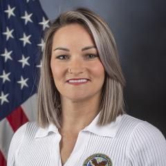 Woman in white shirt with US logo, smiling in front of American flag.