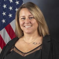 Blonde woman in black top and jacket smiling with American flag behind her.