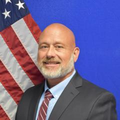 Man in business suit with American flag in background, smiling.