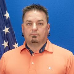 Man with short hair and beard wearing orange polo shirt in front of a blue flag.