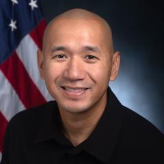 Headshot of a man smiling with an American flag in the background.