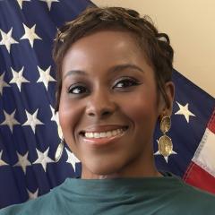 Woman with short hair and green shirt smiling in front of American flag.