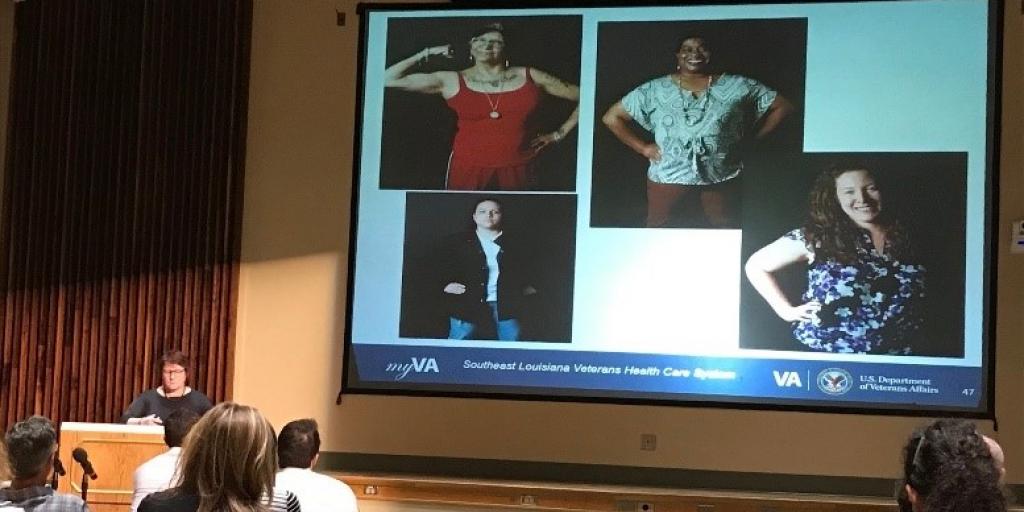 A female presenter shows images on a screen to a room of participants. 