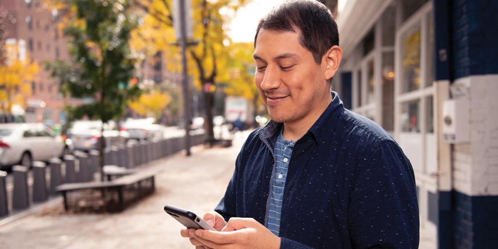 photo of a person with short dark hair, wearing a blue jacket and holding a cell phone with a streetscape in the background