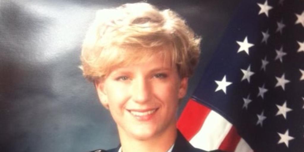 Photo of U.S. Air Force Officer Rebecca Erwin smiling in her Air Force Academy uniform with the American flag in the background. Erwin is a white female with shirt, blonde hair