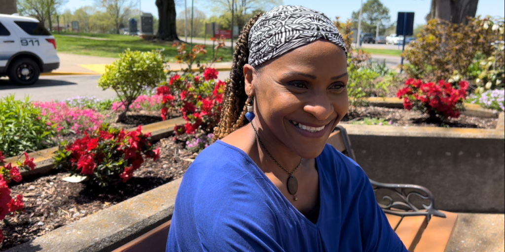 Black female Veteran wearing a blue shirt sits outside in the courtyard next to magenta flowers.