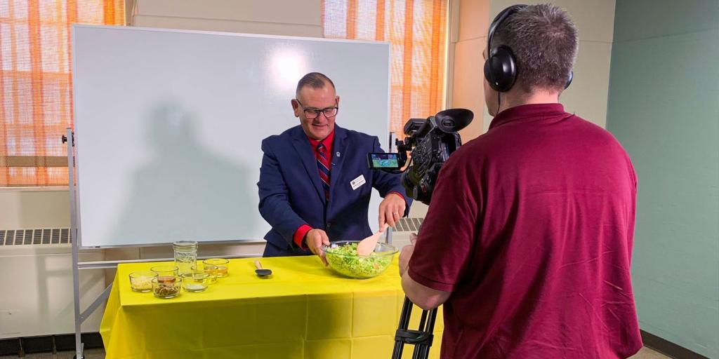 A man stands behind a table while stirs a glass bowl of chopped Brussels sprouts, while being filmed.