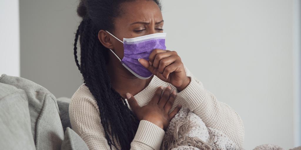 A woman of color wearing a surgical mask and coughing