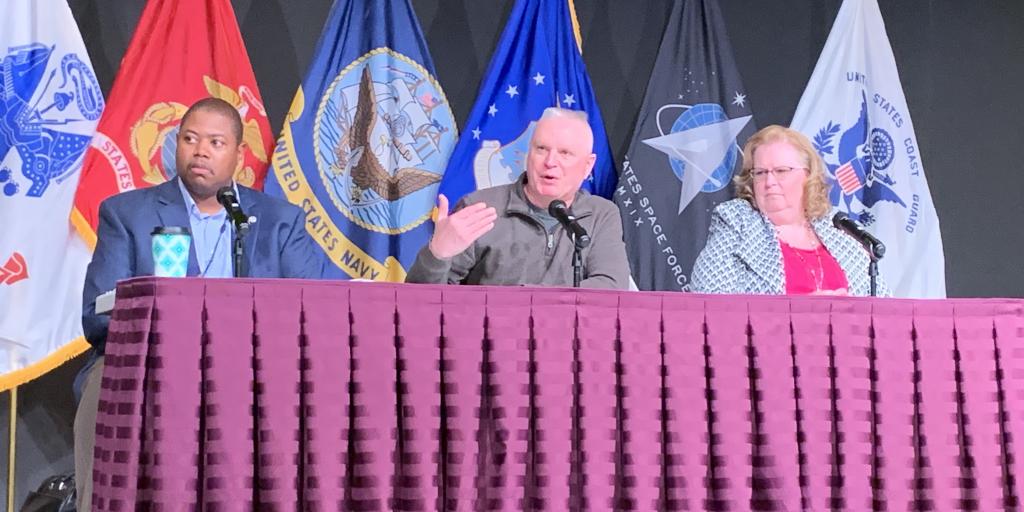 Three panelists on a stage with military flags behind them