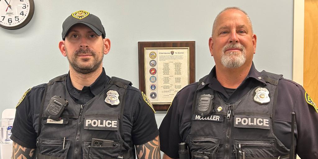 Two male police officers from the Bay Pines VA Healthcare System's Police Service are pictured here with their body cameras. They are standing shoulder to shoulder, in front of a wall in the police station. The gentleman on the left is wearing a dark navy ball cap.