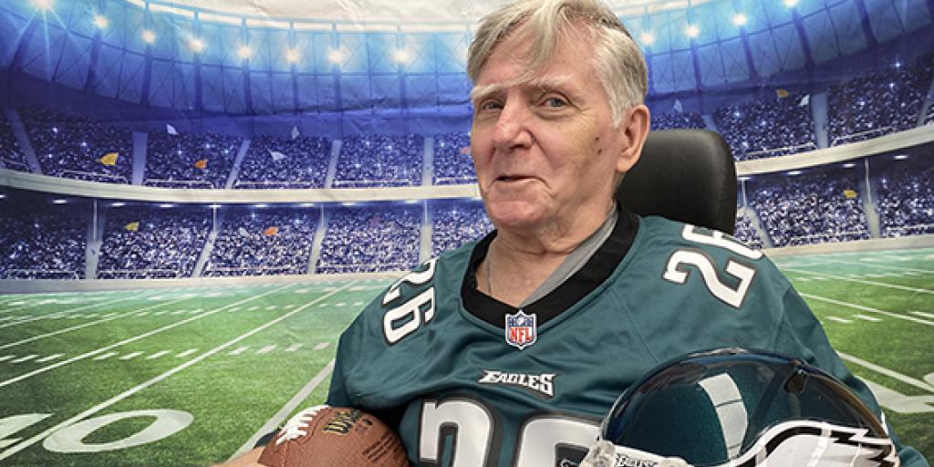 A Veteran in an Eagles jersey is in front of a backdrop of a football field holding a football and a helmet. 