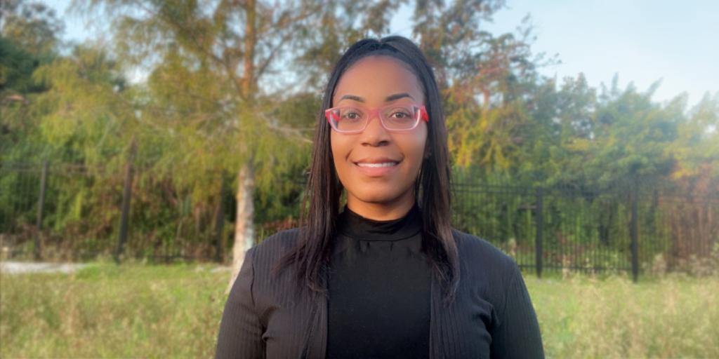 A smiling woman stands outdoors during the daytime, in an open wooded area. She is wearing all black, glasses, and has her hair tied up.