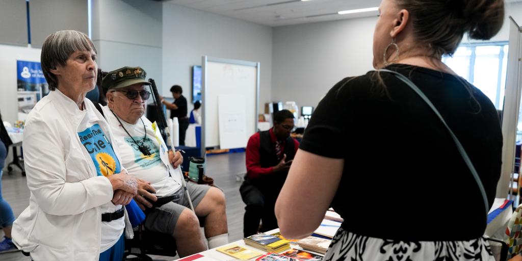 Image of a man in a wheel chair, talking to a woman across a table in a convention center environment.