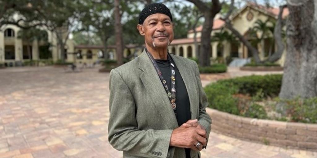A man stands outside, smiling, during the daytime with a blazer and his hands clasped casually in front of him.