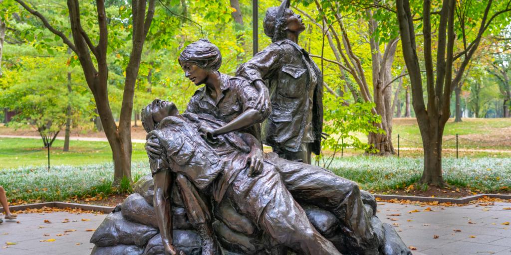 Statue featuring seated woman with man draped across her lap; second woman standing, looking upward.