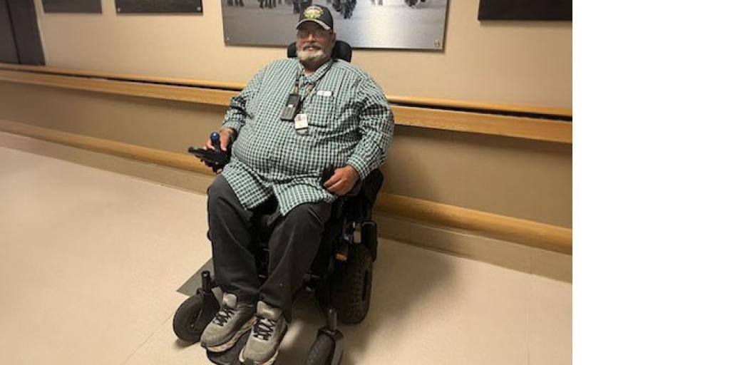 Man sits in wheelchair in hallway, photos behind him