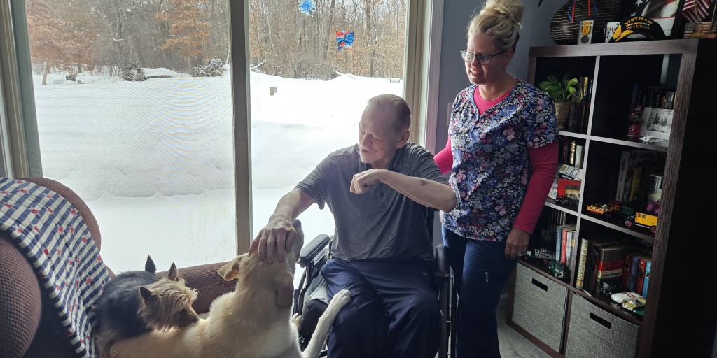 man in wheelchair petting a dog that is sitting on a recliner. A nurse is watching and smiling in the background.