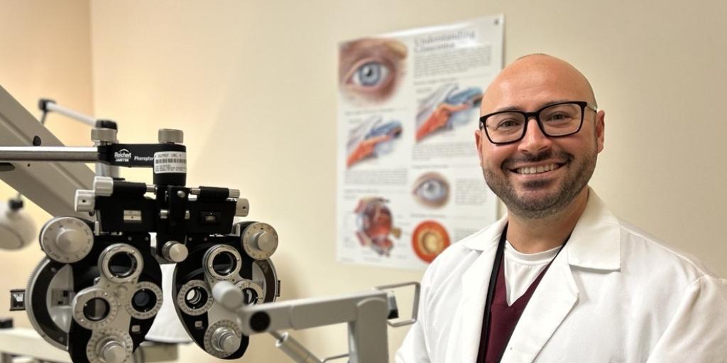 A man in a white lab coat stands smiling in an optical exam room.