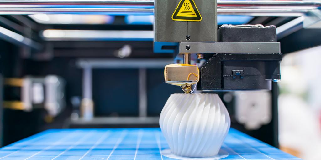 A 3D Printer creates a white spiral resin device on a blue mat with white 1 inch grid squares.