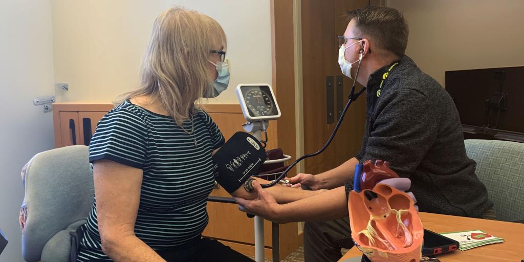 Woman with medical mask sits with blood pressure cuff on her arm. Man, also with mask, observes the pressure gauge.