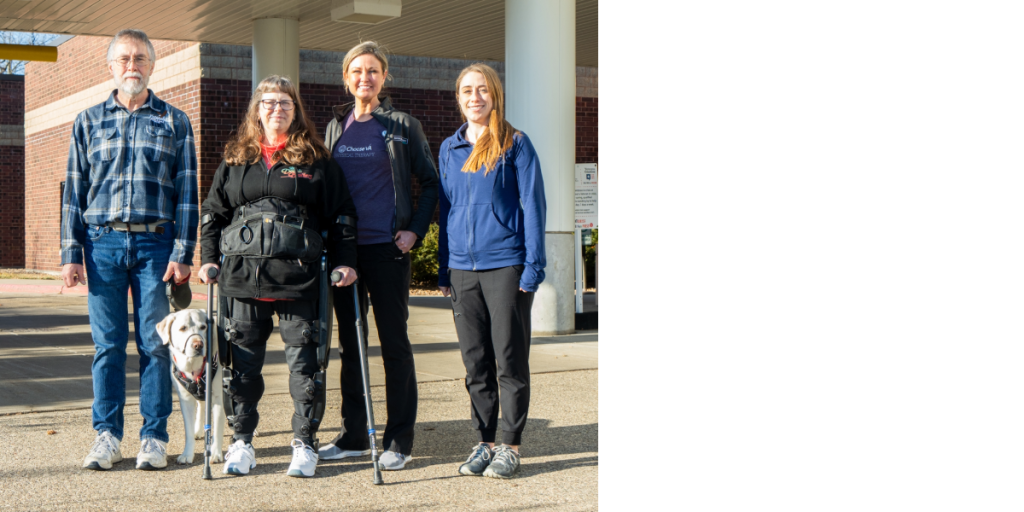four people, one wearing an exoskeleton, and a dog are standing in a row in front of a beige building