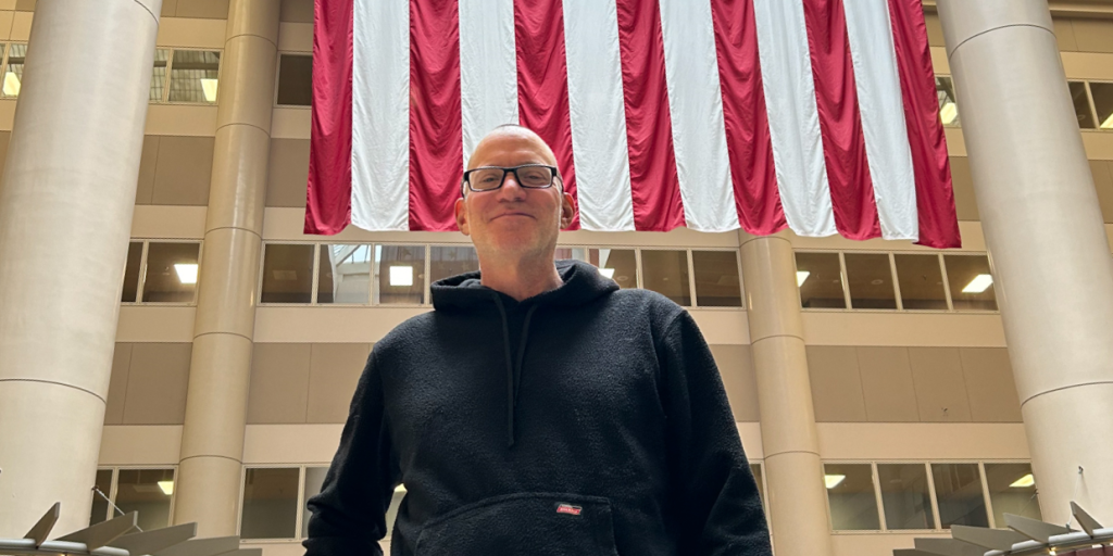 person with short hair and glasses wearing a dark sweatshirt, large columns and the American flag in the background