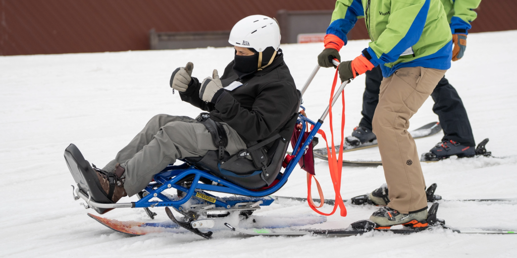 Three people skiing on a snow covered hill. All are wearing ski gear. Person in front is using adaptive skis.