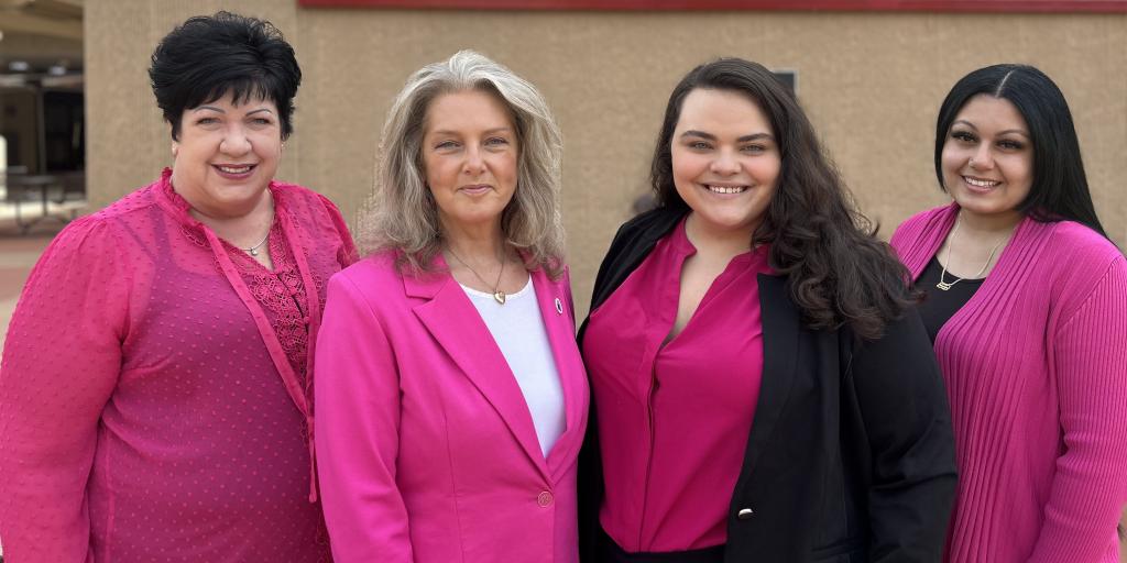 Four women wearing the same shade of pink, stand together outside and smiling. Behind them is a mural of a woman service member holding her young son.