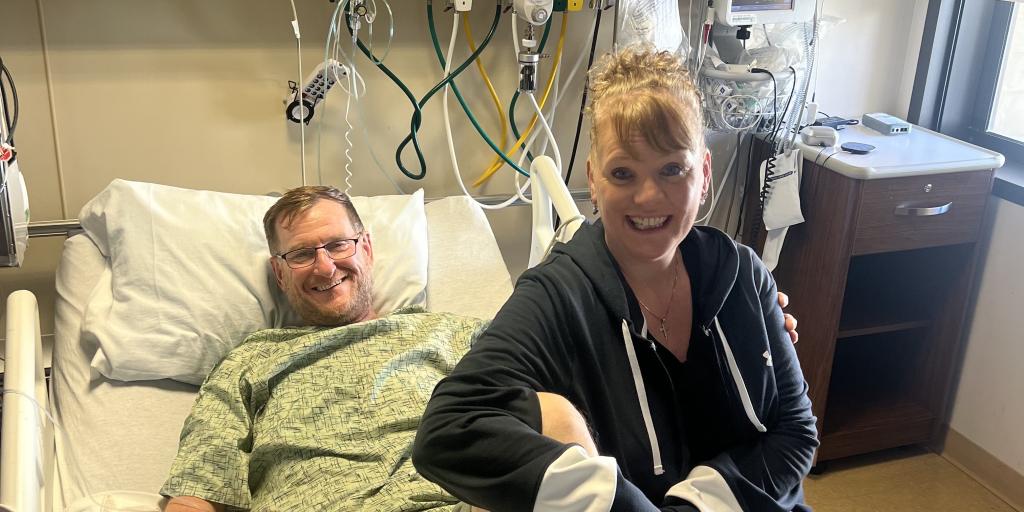 Male, above leg amputee lies in hospital bed with his wife immediately to his right perched on the edge of the bed, smiling as they pose for a photo. 