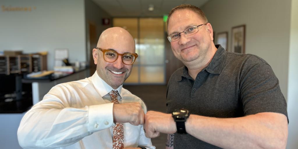Two men, dressed in business casual attire, stand next to each other in a waiting room. They are fist bumping.